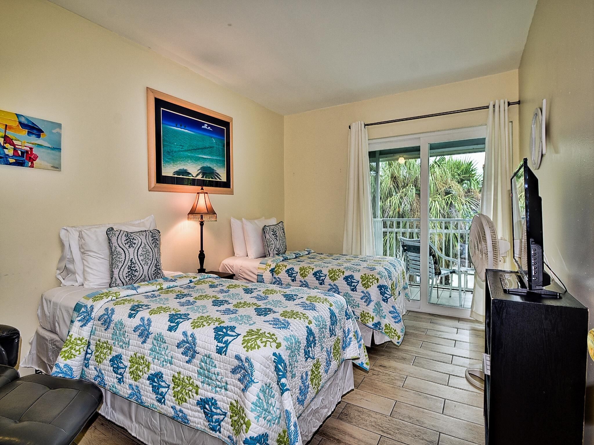 Habitación de hotel con dos camas, un balcón con vistas a palmeras, un cuadro de una playa y un televisor sobre un mueble.