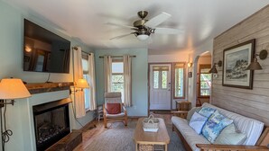 A living room with a fireplace, a ceiling fan, a sofa, a chair, a coffee table, and a television. - Follow The Heron: Fantastic home with lots of decks. (Ocracoke)