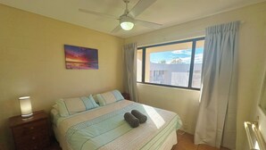 Une chambre à coucher comprenant un lit, une table de chevet, un ventilateur de plafond, une fenêtre avec des rideaux et un tableau accroché au mur.