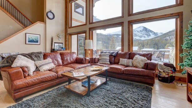 A spacious living room with a large brown leather sectional sofa, a wooden coffee table, and a view of snow-capped mountains through large windows.