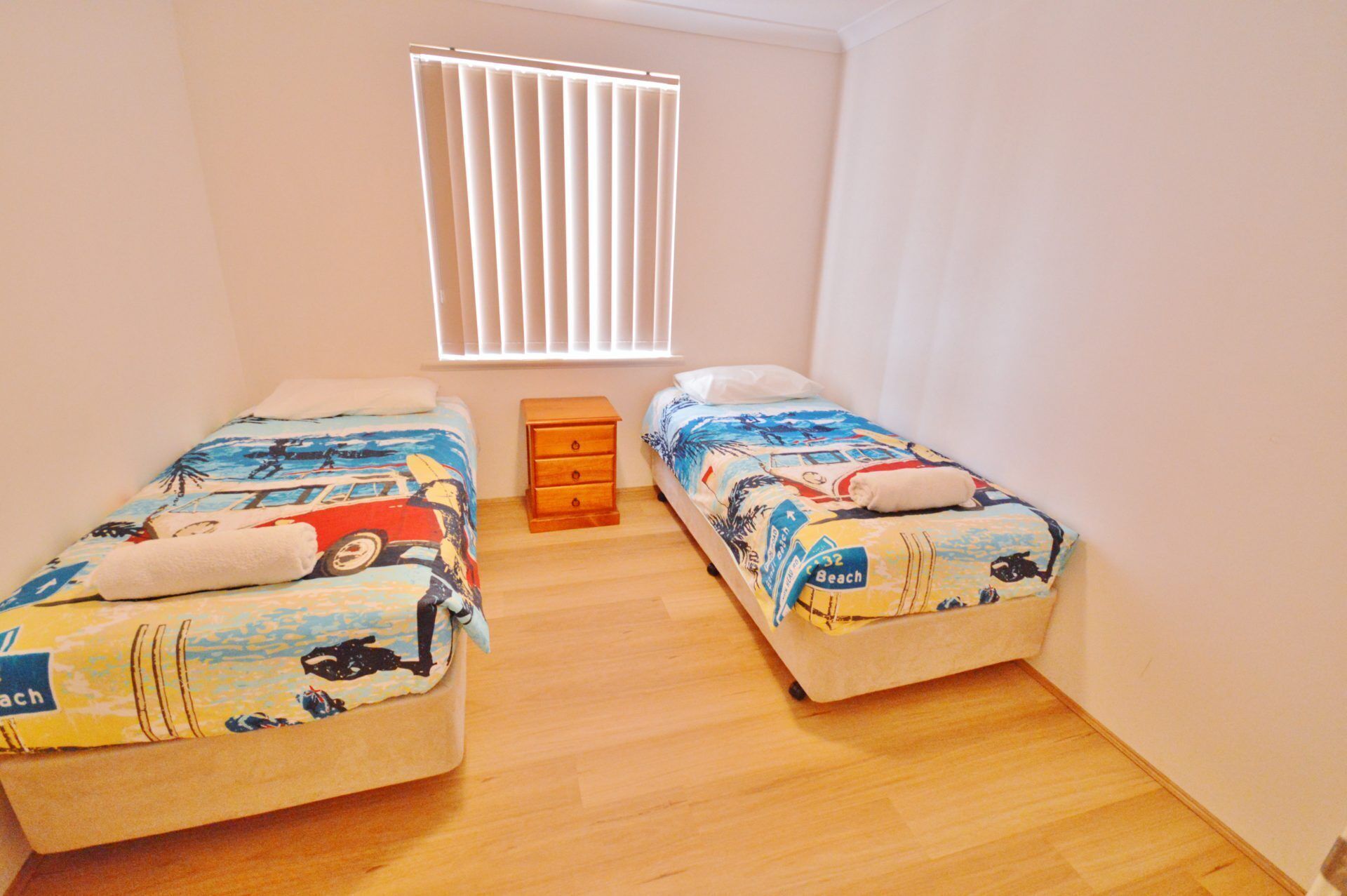 Une petite chambre propre, avec deux lits simples, une commode en bois et une fenêtre avec des stores.