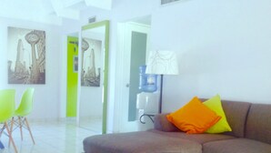 Una sala de estar con un sofá, una mesa de centro de vidrio y una lámpara. Hay dos sillones y una puerta que da a otra habitación.