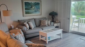 Een woonkamer met een grijze bank, een witte salontafel, een plafondventilator en een groot raam met uitzicht op groen.