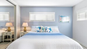 A bedroom with a bed, two bedside tables with lamps, a window with blinds, and a wall-mounted picture.