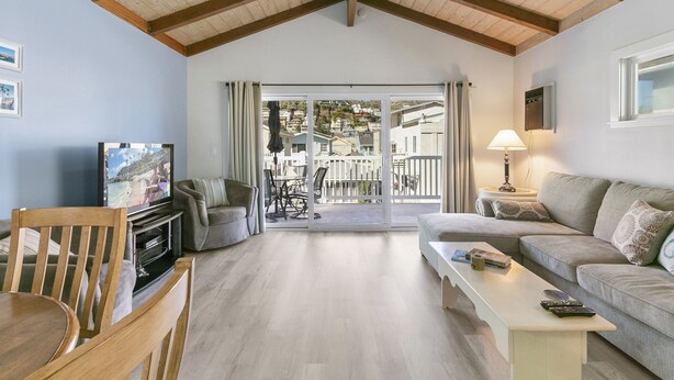 A living room with a wooden ceiling, a television, a sofa, a coffee table, and a balcony with a view of houses.