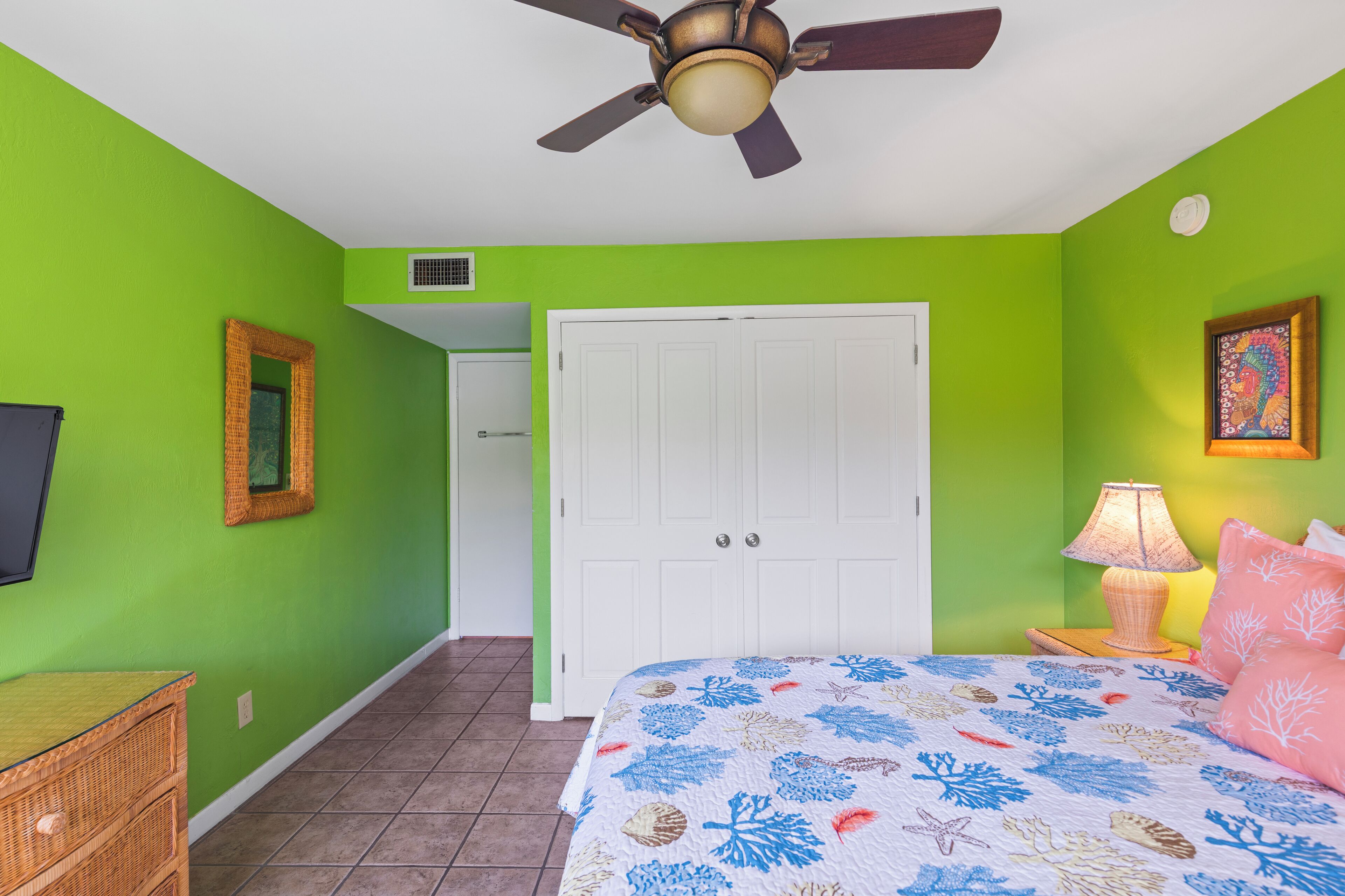 A bedroom with a bed, a ceiling fan, a door, a wall-mounted picture, a bedside lamp, and a wicker nightstand.