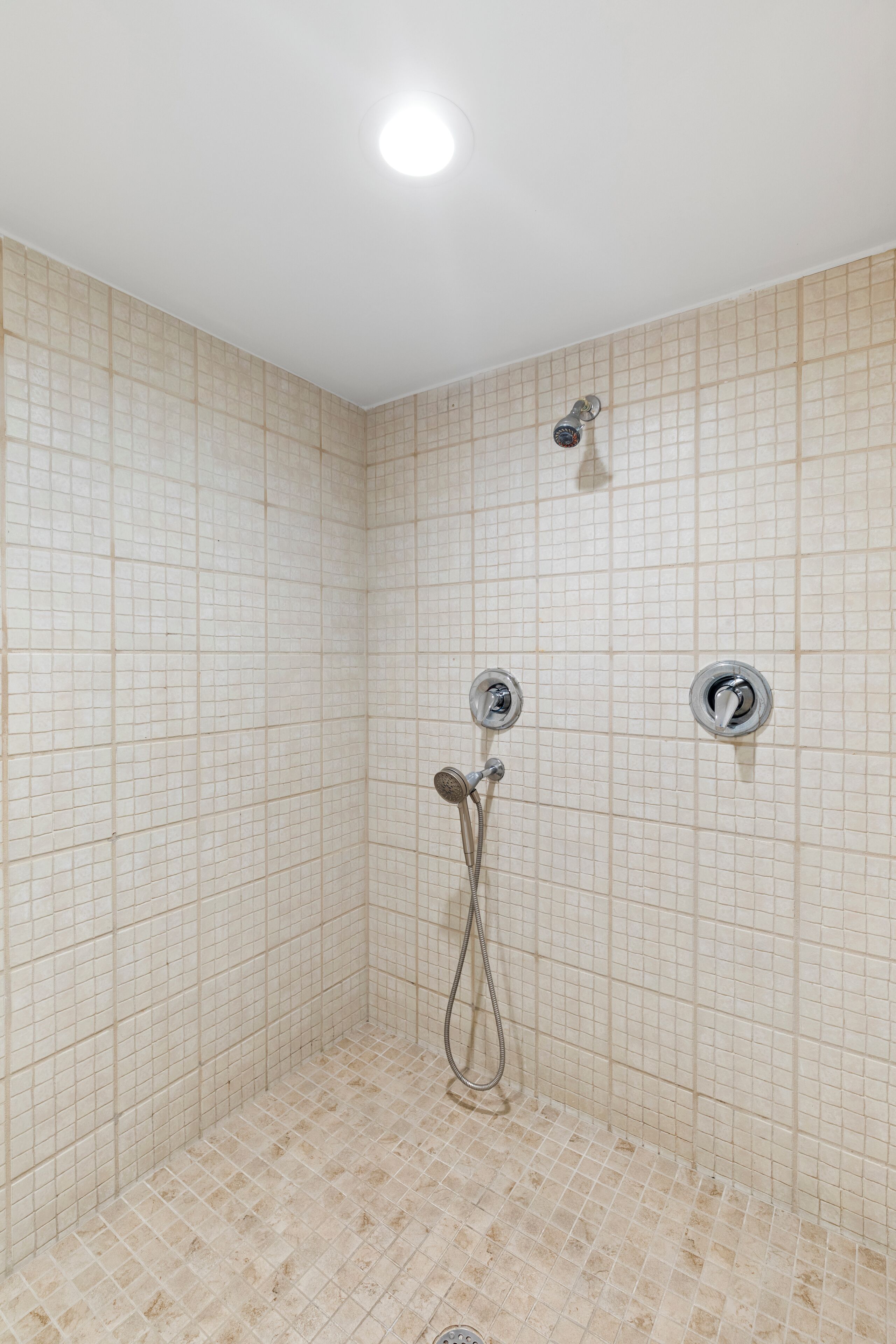 A tiled shower with a handheld showerhead and two wall-mounted faucets.