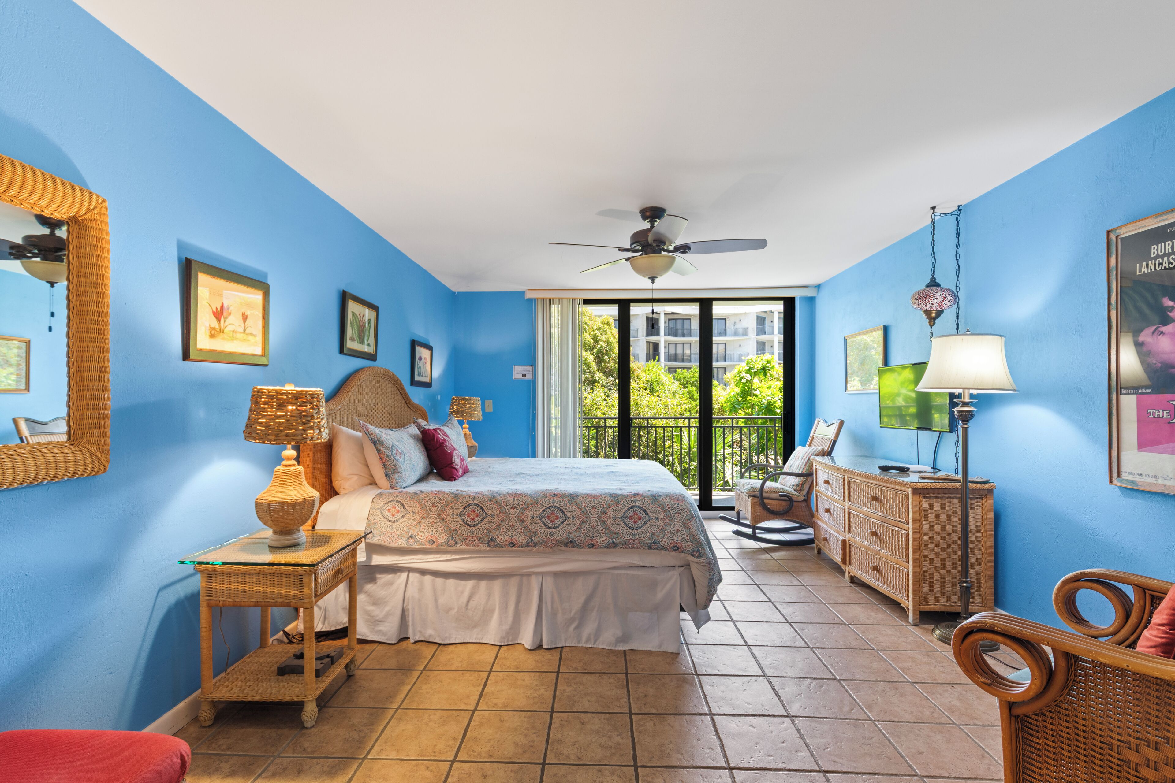 A bedroom with a bed, a ceiling fan, a dresser, a chair, and a balcony with a view of greenery.