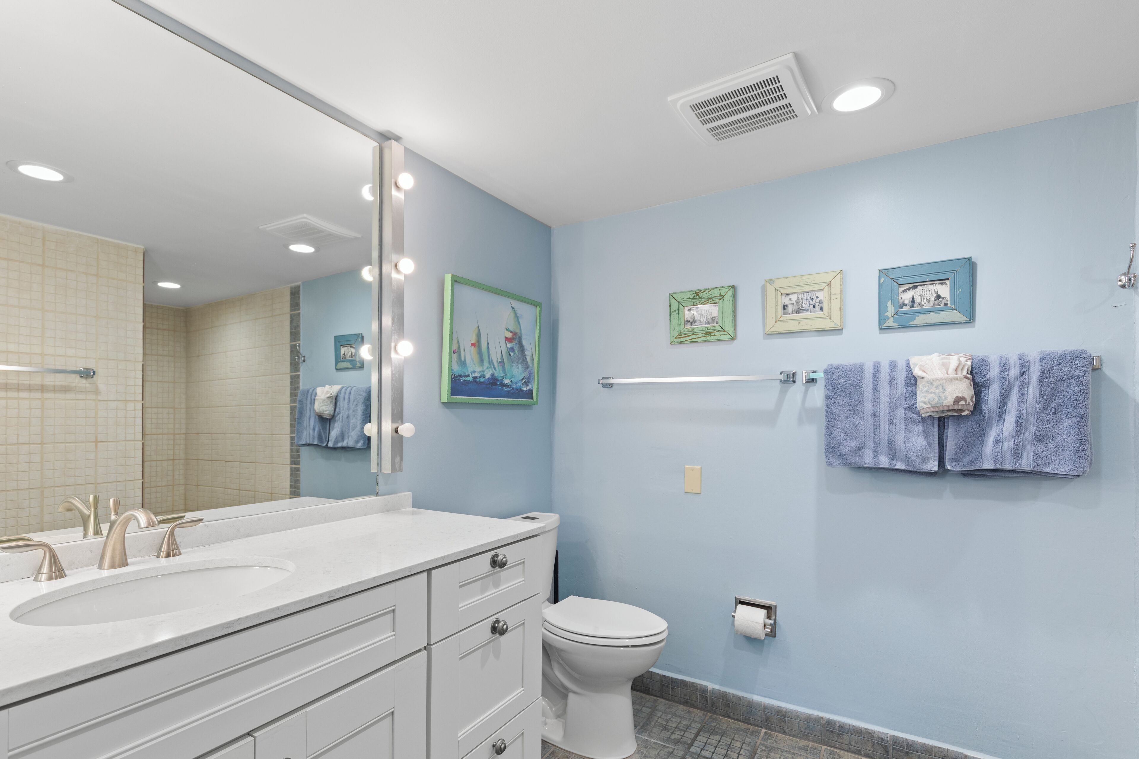 A bathroom with a double sink, white vanity, and a toilet. There are framed pictures on the wall and a towel rack with towels.