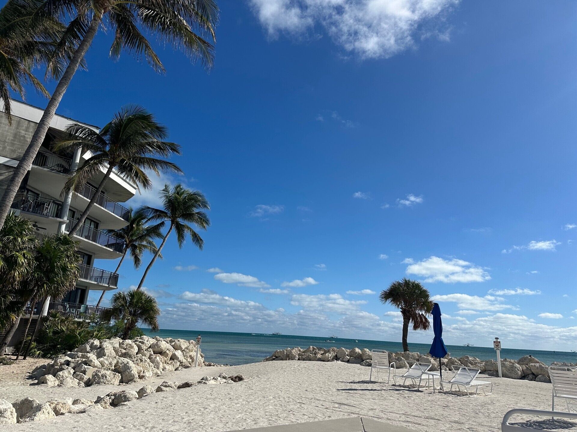 A beach with white sand, palm trees, and a building with balconies overlooking the ocean.