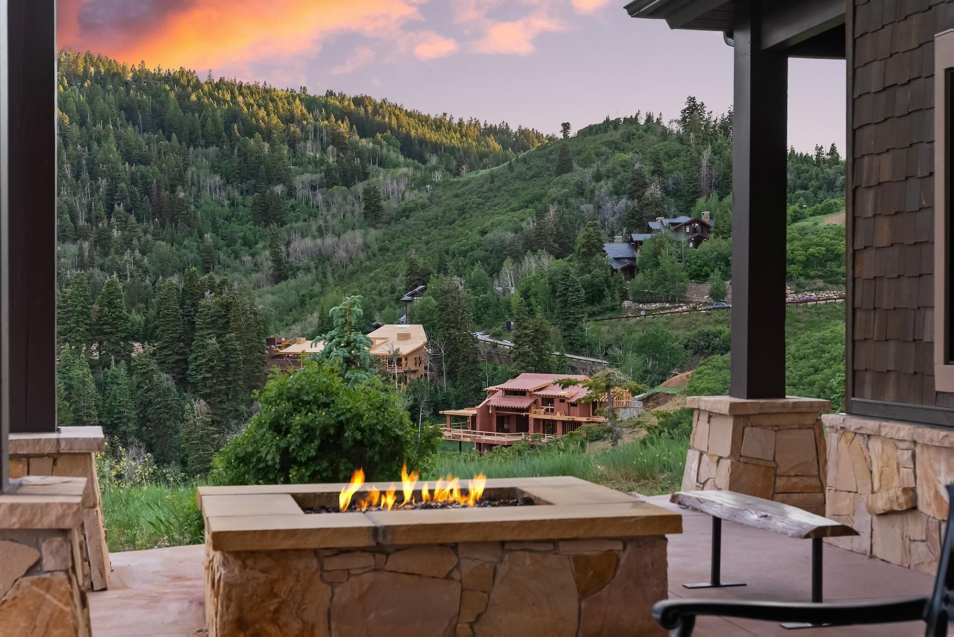 A stone fire pit with flames, surrounded by stone walls and a bench, overlooking a forested hillside with several buildings.