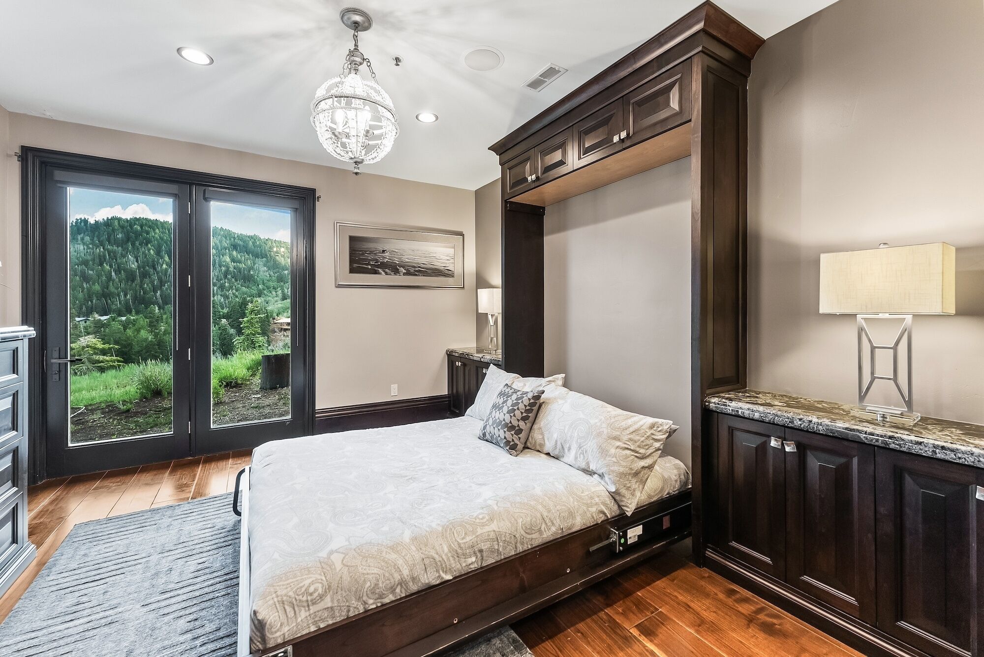 A bedroom with a large bed, a chandelier, a view of a forest through a sliding door, and dark wood paneling.