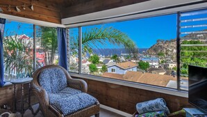 A room with a large window offering a view of a coastal town, a wicker chair, and a small table with a sign that reads "BEACH". - Cozy 2 Bedroom Home with GOLF CART, Stunning View, Balcony, Grill, Wifi - 124 Hi (Avalon)
