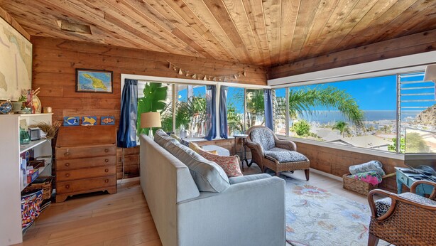 A spacious living room with a wooden ceiling, large windows offering a view of the ocean, a sofa, a chair, and a small table. - Cozy 2 Bedroom Home with GOLF CART, Stunning View, Balcony, Grill, Wifi - 124 Hi (Avalon)