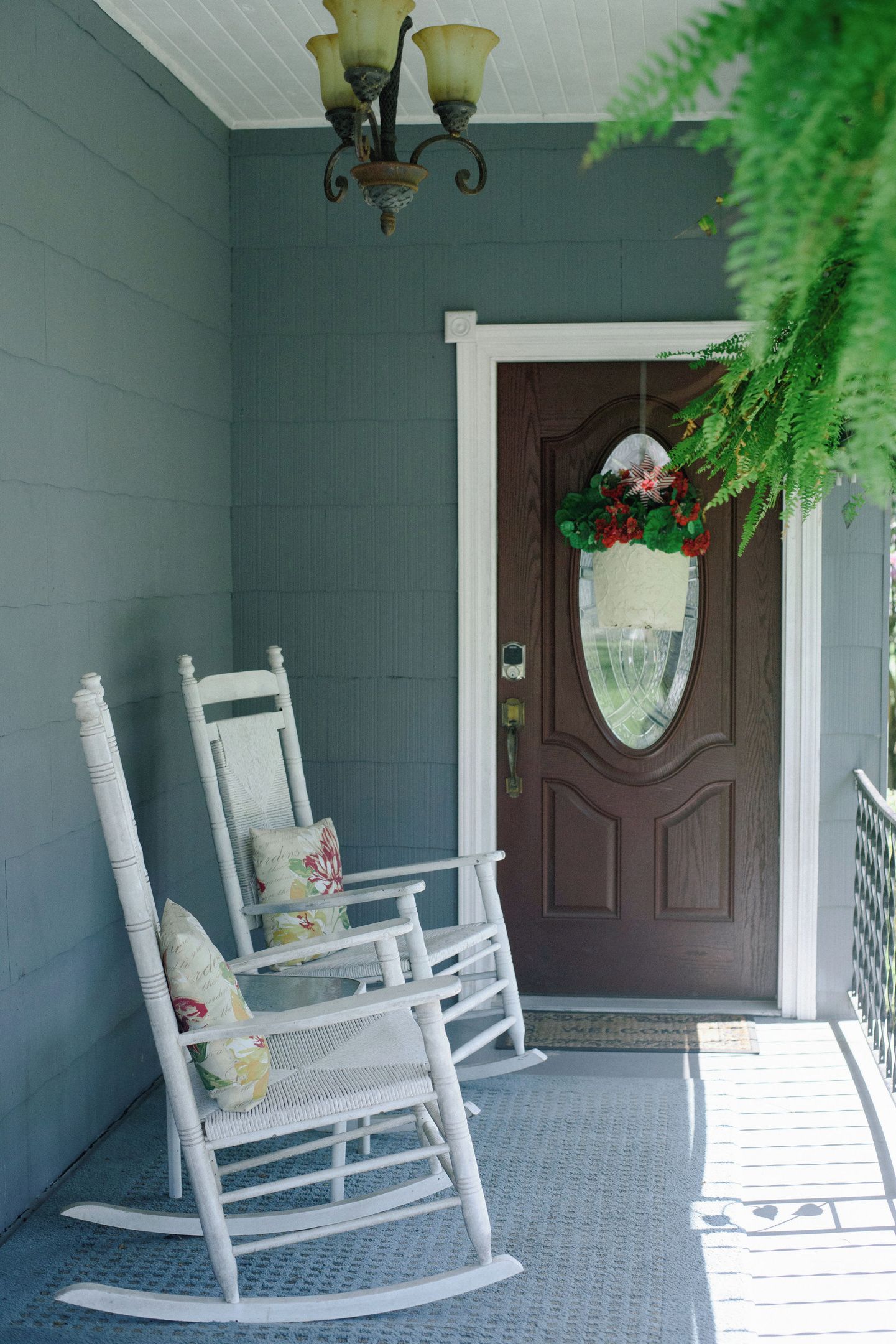 A porch with two rocking chairs, a brown door with a decorative glass panel, and a hanging plant.
