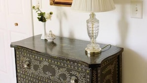 A dark wooden dresser with decorative patterns and a gold-framed mirror above it. A white vase with flowers and a white lamp with a pleated shade are on the dresser. - The Original Crafter's Cottage Convenient to Savannah (Bloomingdale)