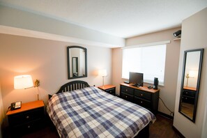 A hotel room with a bed, bedside tables, a desk with a computer, a mirror, and a window with blinds. - Marketplace Lodge 333 (Whistler)