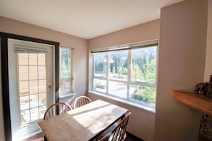 Una habitación con una mesa de comedor, sillas, un ventanal con vistas a árboles y un edificio, y una puerta con persianas.