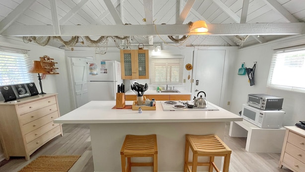 Une cuisine moderne avec des meubles blancs, un îlot central, des tabourets en bois et un plafond avec des poutres apparentes.