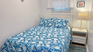 Une chambre à coucher comprenant un lit, une table de chevet, une lampe, une fenêtre avec des stores et un tableau accroché au mur.