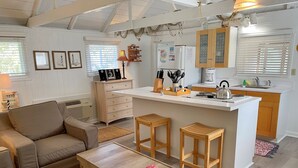 Un salon chaleureux avec un canapé, une table basse et des tabourets en bois. On aperçoit à l’arrière-plan un coin cuisine avec un plan de travail blanc et des placards en bois.