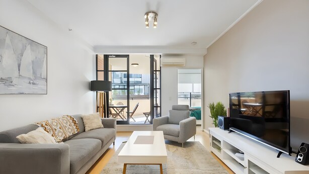 Una sala de estar moderna con un sofá gris, una mesa de centro blanca, un sillón gris, un televisor de pantalla plana y un ventanal con vistas al exterior.