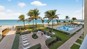 Zwembad van een resort met palmbomen, ligbedden en uitzicht op strand en zee.