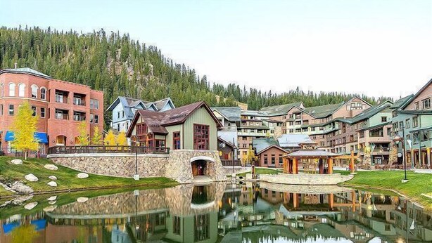 Un resort junto al lago con un puente de piedra, edificios reflejados y un bosque de fondo.