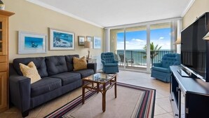 Een woonkamer met een donkere bank, twee fauteuils, een glazen salontafel en een flatscreen televisie. Er hangen prestatiekaders aan de muur en is er een balkon met uitzicht.
