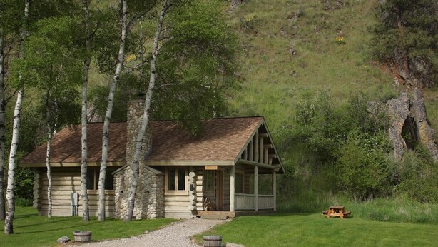 A log cabin with a stone chimney, surrounded by trees and a picnic table. - Aspen Cabin at Rye Creek Lodge (Darby)