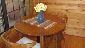 A wooden dining table with a vase of flowers, a woven placemat, and a wooden chair with a cushion. -  Romantic Log Cabin On Creek And 100 Acres, Hot Tub, Wifi, Gas Grill, Hiking (Hot Springs)