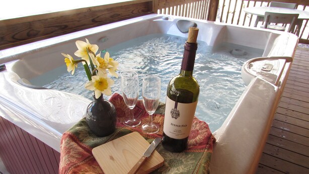 A hot tub with a bottle of wine, glasses, and a vase with flowers on a wooden deck. -  Romantic Log Cabin On Creek And 100 Acres, Hot Tub, Wifi, Gas Grill, Hiking (Hot Springs)