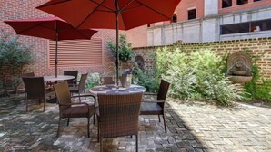 An outdoor patio with wicker furniture and red umbrellas, brick walls, and a stone fountain. - 1bd, 1.5ba Historic District and River Front-Walk to Everything! (Savannah)