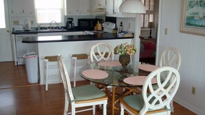 A dining area with a glass table surrounded by white chairs with green cushions, a white trash bin, and a kitchen in the background. - Shell House  - Less than a block to beach access and recently updated! (Sunset Beach)