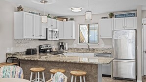 Una cocina moderna con gabinetes blancos, encimera de granito, refrigerador de acero inoxidable y una barra con taburetes.