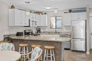 A modern kitchen with white cabinets, a granite countertop, a stainless steel refrigerator, and a bar area with stools.