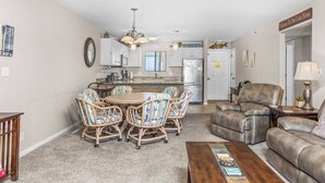 A living room with a dining table, chairs, a sofa, and a coffee table. There is a kitchen area with cabinets and appliances in the background. - Lazy Days V-01 - Five Bedroom Apartment, Sleeps 12 (Osage Beach)