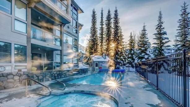 Une piscine avec un jacuzzi, entourée d’arbres couverts de neige et d’un bâtiment doté de balcons.