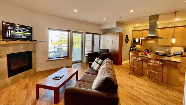 A modern living room with a sofa, a coffee table, and a TV on a stand. There is a kitchen area with a stove, sink, and bar stools. Large windows provide natural light and a view of the outdoors.