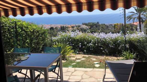 Un porche cubierto con una mesa y sillas, con vista a un pueblo costero y un jardín con flores moradas.