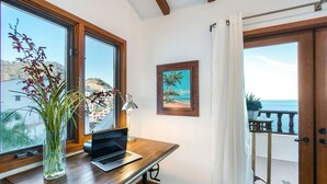 Ein Zimmer mit Schreibtisch aus Holz, Laptop und einer Pflanzvase. An der Wand hängt ein Bild vom Strand, und ein Balkon mit Meerblick ist zu sehen.