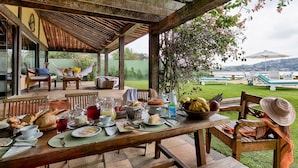 Un área de comedor al aire libre cubierta, con una mesa dispuesta para el desayuno, incluyendo pan, frutas y bebidas.