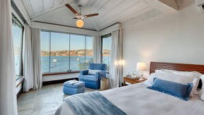 Una habitación de hotel con una cama grande, un sillón azul, una mesita de noche de madera y vista al mar.