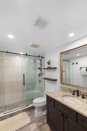 Une salle de bain moderne avec une douche à parois de verre, des toilettes blanches, un lavabo beige doté d’un plan de toilette à motifs, et un grand miroir avec un cadre en bois.