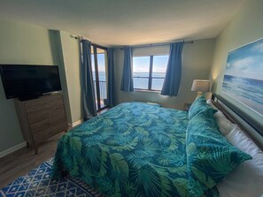 Ein Schlafzimmer mit einem Bett, einem Fernseher auf einer Kommode, einem Fenster mit Vorhängen und einem Bild von einem Strand.