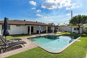 Une piscine résidentielle avec un espace patio, un coin salon couvert et un panier de basketball.