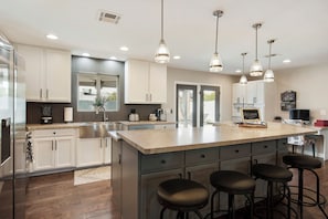 Una cocina moderna con armarios blancos, una isla grande con taburetes de bar, electrodomésticos de acero inoxidable e iluminación colgante.