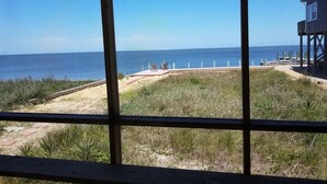 Blick auf einen Strand durch ein Fenster mit Holzbalkon, ein Sandweg und eine Holzkonstruktion mit Treppen zum Strand.