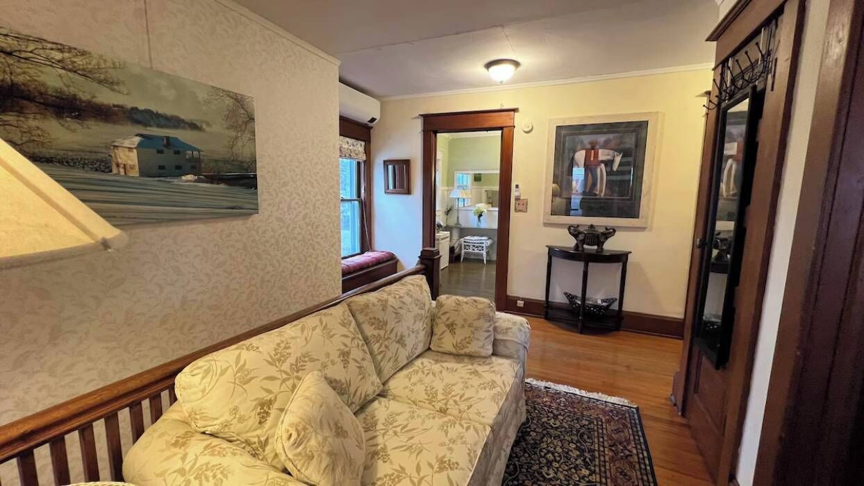 A living room with a floral sofa, a wooden coffee table, and a patterned rug. There is a painting on the wall and a mirror above the coffee table.