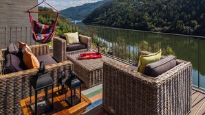 Un balcon agrémenté de meubles en osier, d’un fauteuil suspendu et offrant une vue sur un lac et des collines boisées.
